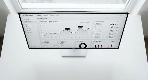 A minimalist, top-down view of a modern enterprise dashboard displayed on a large widescreen monitor. The UI features clean, flat surfaces in light grey and white, with simple bar charts and line graphs in deep black. Red dot indicators denote 'live' syncing across multiple locations. The surrounding desk space is completely empty and white, emphasizing a clean corporate modernism style.